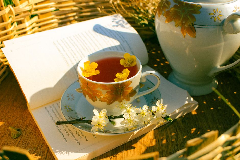 A serene tea setup with a floral cup, teapot, and open book amidst sunlight and blossoms.