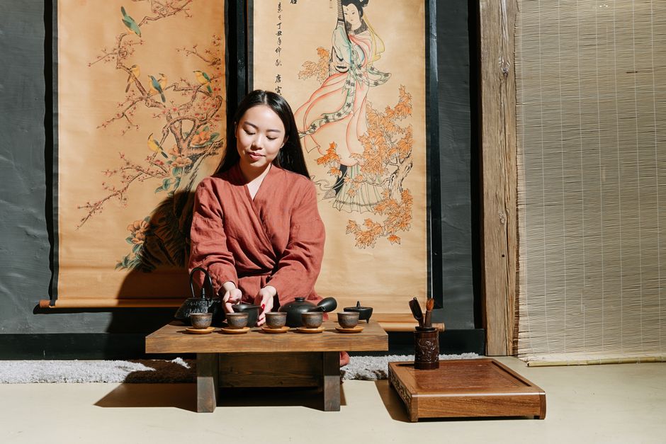 Asian woman performing traditional tea ceremony indoors with decorative scrolls.